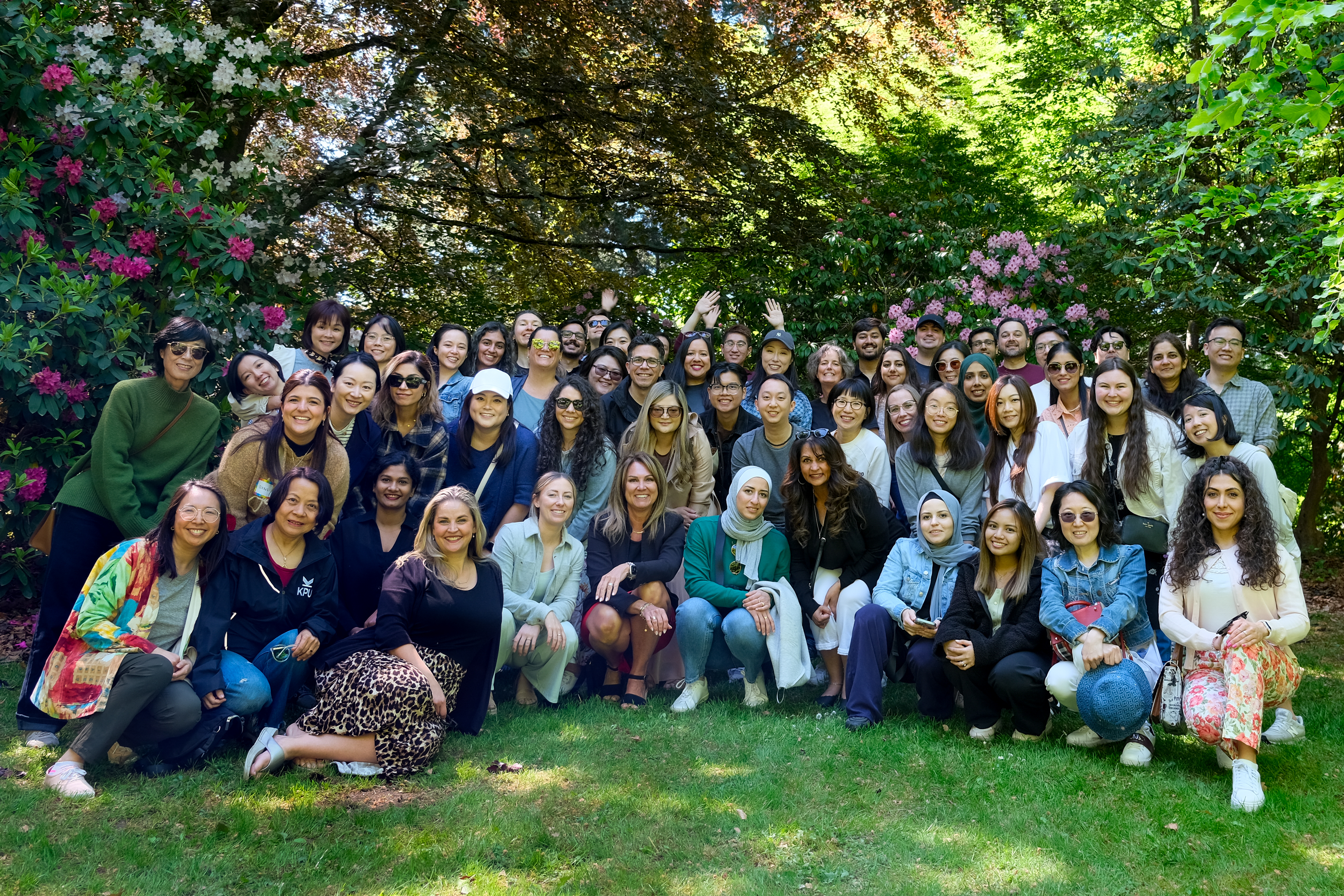 Photo of KPU International staff in a group, standing outside on a grassy lawn and behind are green bushes with flowers and large leaves. 