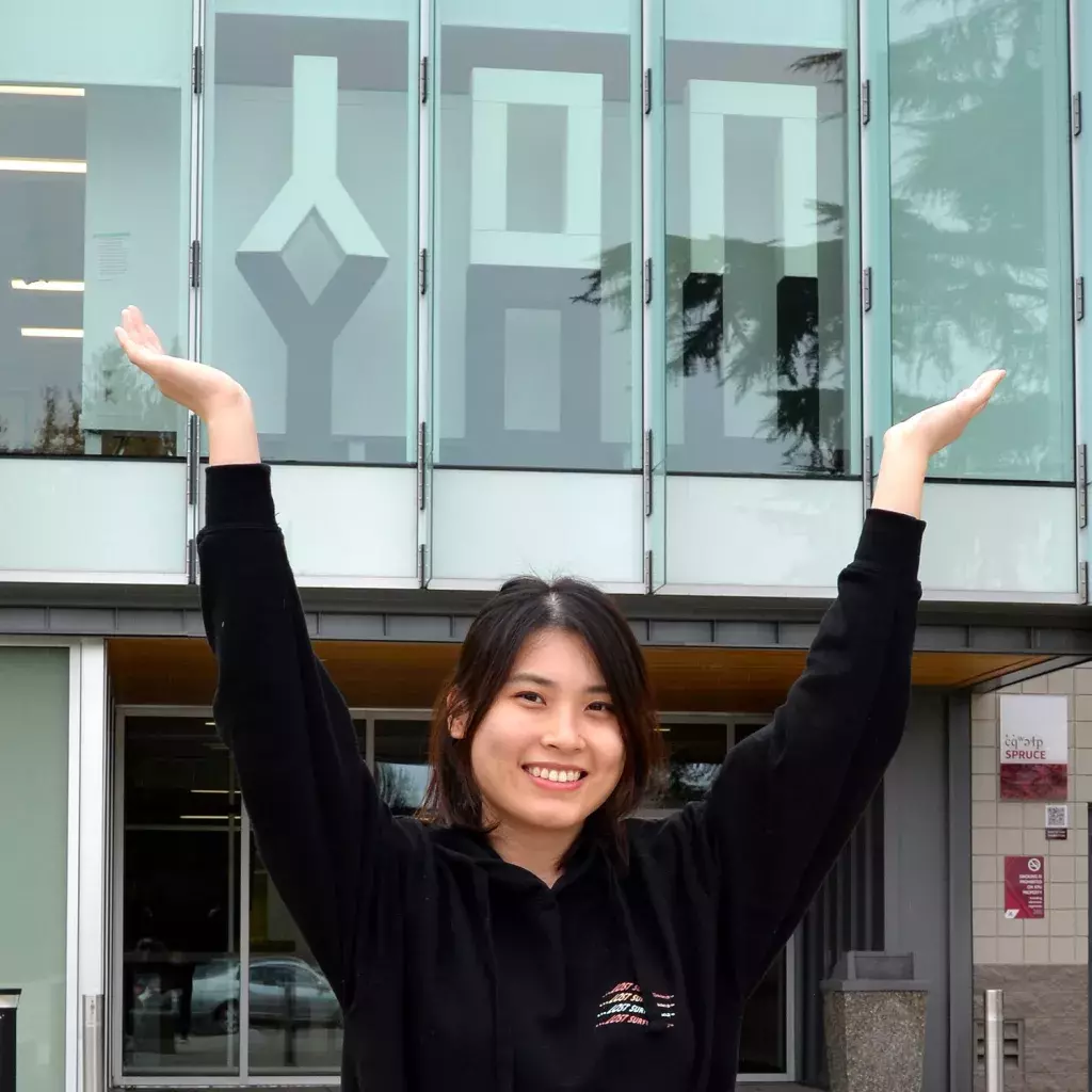 Artist Winnie Hui raises her arms, creating a Y-shape framing the word you in large letters on the first floor of the glass building behind her
