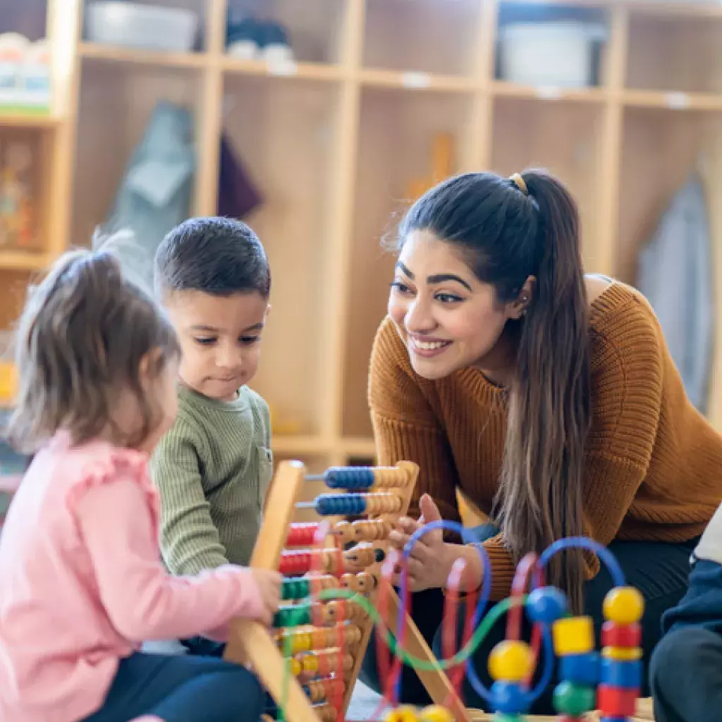 A female Kindergarten teacher of Middle Eastern decent, sits on the floor with students as they play with various toys and engage in different activities. 