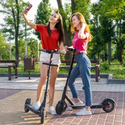 two women outdoors with electric scooters one is holding up a phone taking a photo